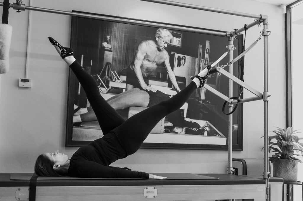 Sonya Rawlinson, a certified Pilates instructor with over a decade of experience, standing in front of a picture of Joseph Pilates in a Pilates studio, wearing comfortable workout attire and demonstrating proper form while performing Pilates on the caddy.